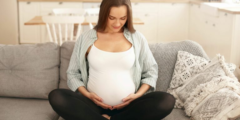 People, lifestyle, pregnancy and anticipation concept. Beautiful young brunette nin month pregnant female sitting barefooted on couch in living room, holding hands on her tummy, feeling quickening